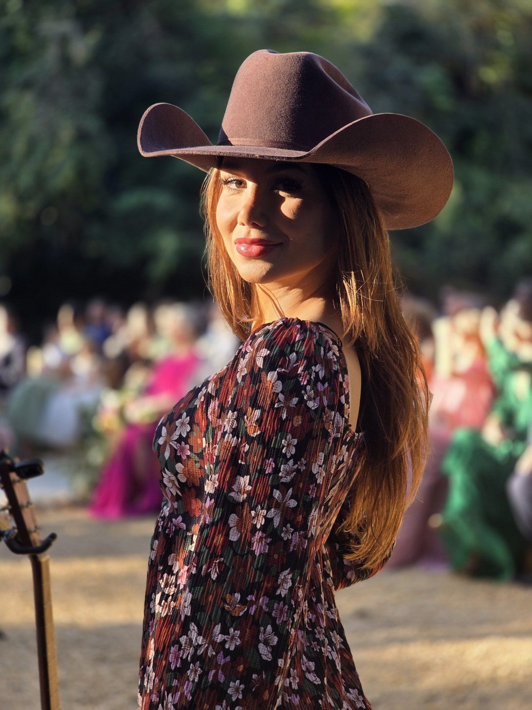 Cantora de chapéu e vestido floral sorrindo na cerimônia ao pôr do sol, com os convidados ao fundo